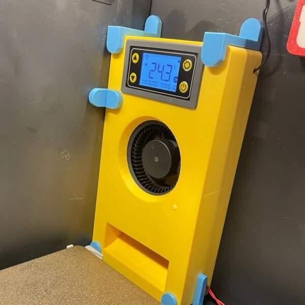 A yellow electronic device with a digital display showing a temperature of 24.3°C, mounted on a wall. The device features a circular fan opening and blue plastic corners for stability.