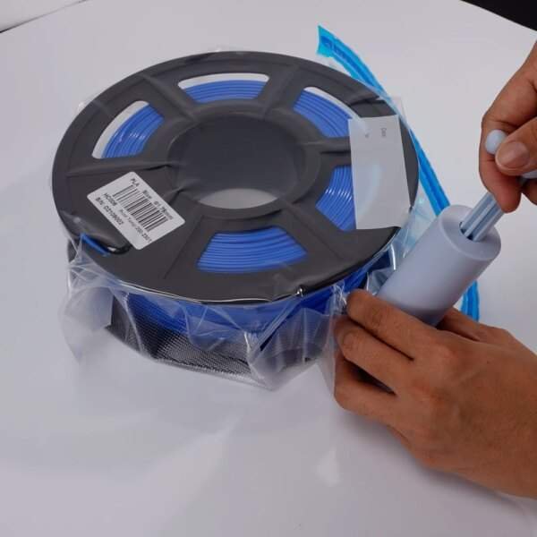 A person is using a vacuum sealer to remove air from a bag containing a spool of blue filament, which is resting on a white surface.