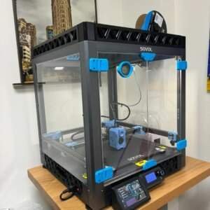 A 3D printer enclosed in a clear plastic case with blue accents sits on a wooden table, with a display screen and control panel in front. In the background, a photograph of a tall building is visible on the wall.