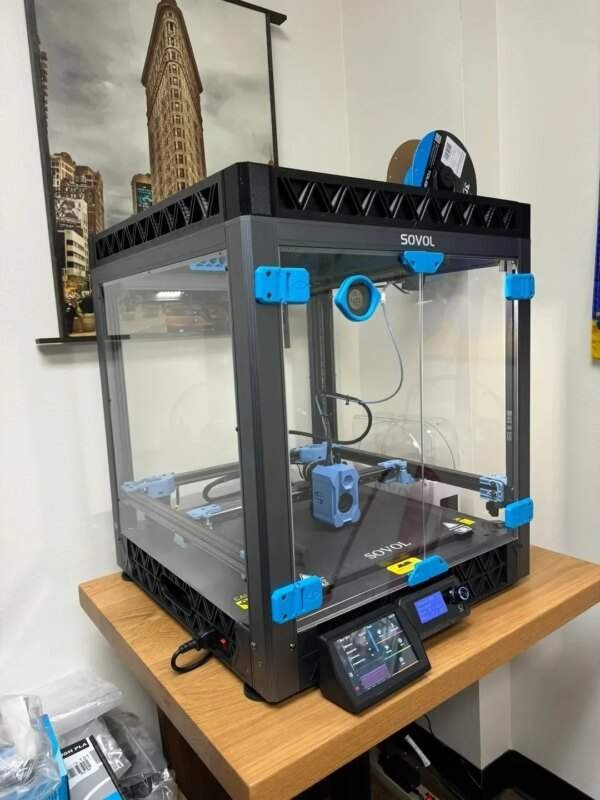 A 3D printer enclosed in a clear plastic case with blue accents sits on a wooden table, with a display screen and control panel in front. In the background, a photograph of a tall building is visible on the wall.
